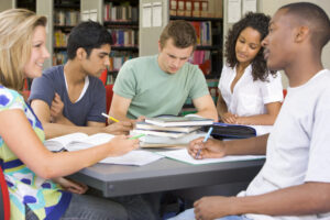 A group of five people seating on a table which is filled with books and notebooks. 