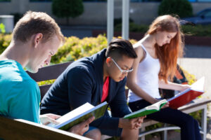 Three students, one woman and two men siting on a bench. They are squinting at the books they are holding.